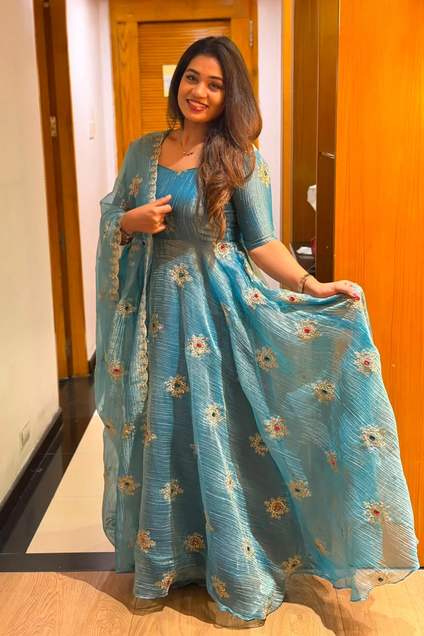 A woman wearing a light blue traditional dress with floral embroidery and a matching dupatta, standing in an indoor hallway with wooden doors.