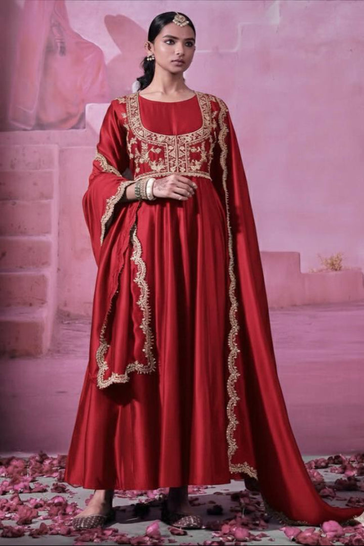 Woman in a red traditional outfit with gold embroidery standing on a pink background with rose petals.