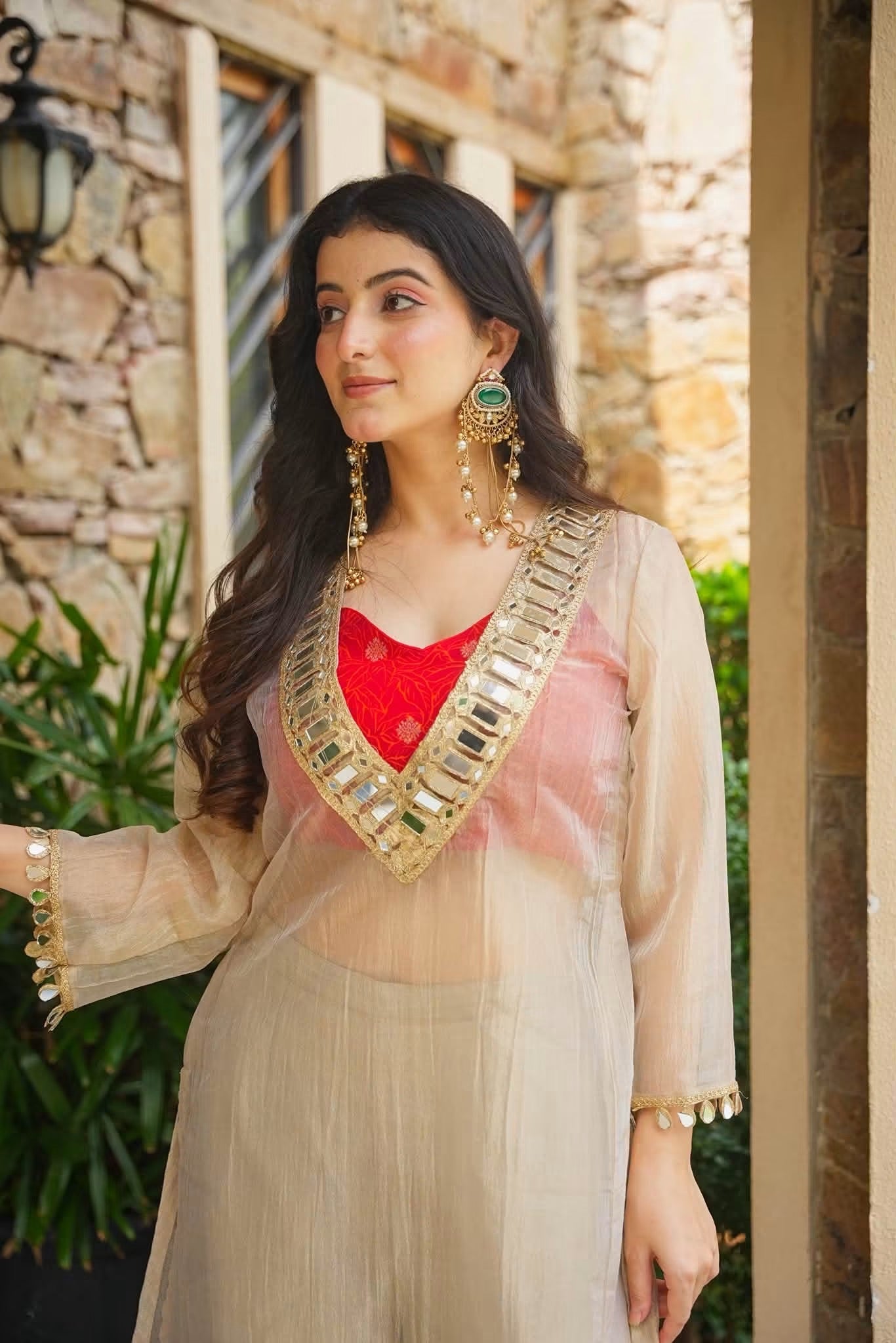 A woman wearing a beige sheer embroidered kurta over a red top, standing outdoors in front of a stone wall.