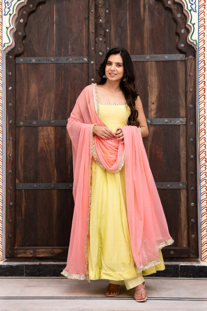 Woman in a yellow dress with a pink dupatta standing in front of a wooden door.