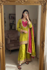 A woman takes a mirror selfie wearing a traditional Indian outfit consisting of a lime green kurta with pink and gold embroidery, matching wide-leg pants, and a bright pink dupatta with gold borders. The photo is taken in front of an ornate gold-framed mirror in a living room setting.