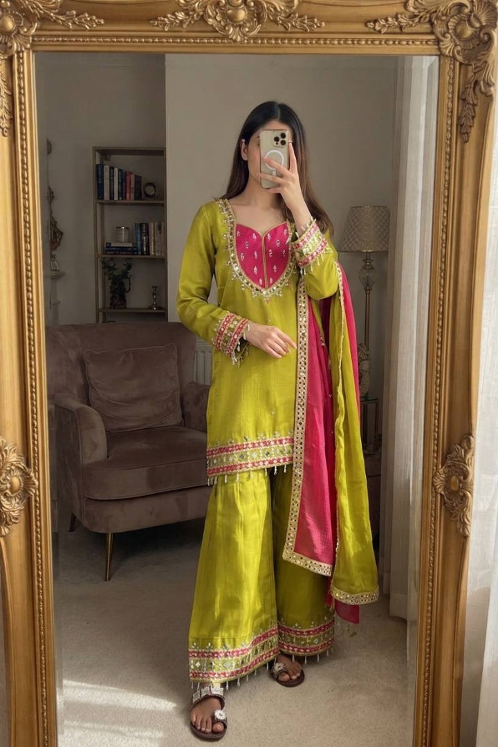 A woman takes a mirror selfie wearing a traditional Indian outfit consisting of a lime green kurta with pink and gold embroidery, matching wide-leg pants, and a bright pink dupatta with gold borders. The photo is taken in front of an ornate gold-framed mirror in a living room setting.