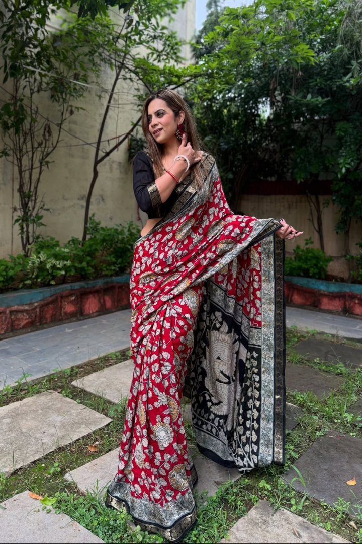 Woman holding a red and black saree in an outdoor setting with greenery.
