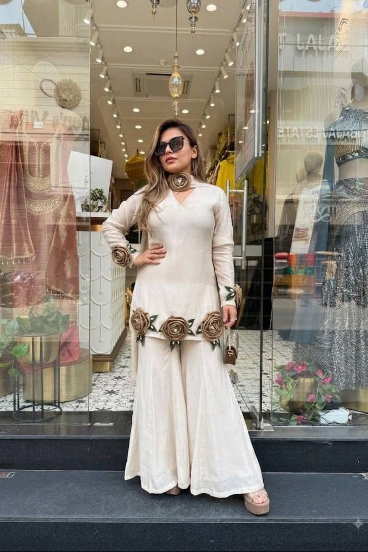 A woman wearing a cream-colored long-sleeve maxi dress with floral embroidery stands in front of a clothing boutique window display.