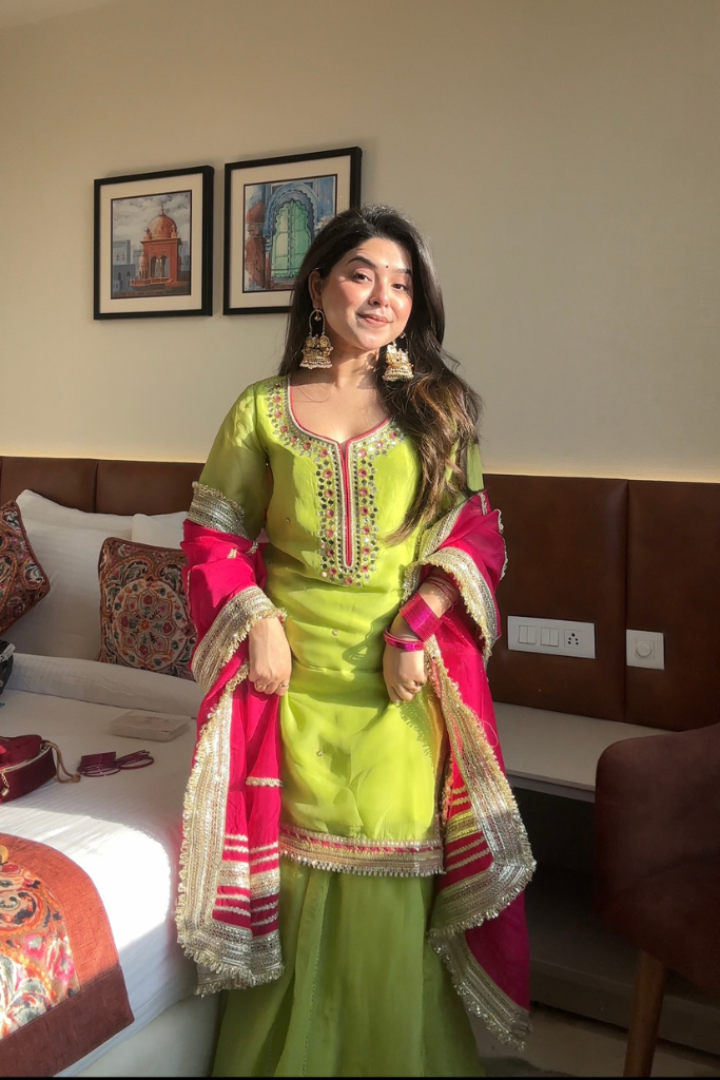 Woman in a green and pink traditional outfit standing in a room with a bed and wall art.