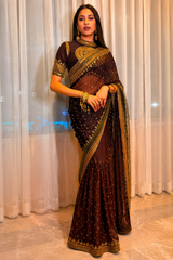 Woman wearing a traditional brown saree with gold accents against a white curtain background