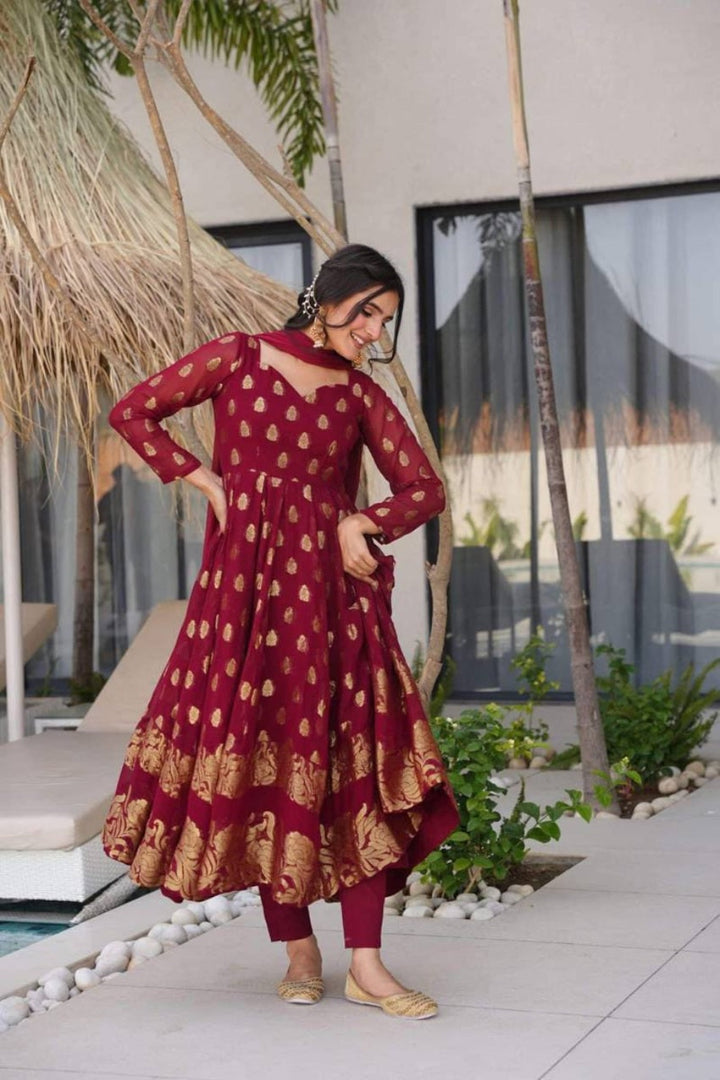 Woman in a maroon and gold traditional outfit standing outdoors with plants and a building in the background.