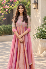 Mrunal Thakur wearing a pink and peach embroidered traditional dress with sheer sleeves, standing outdoors in front of a beige wall with greenery and flowers.