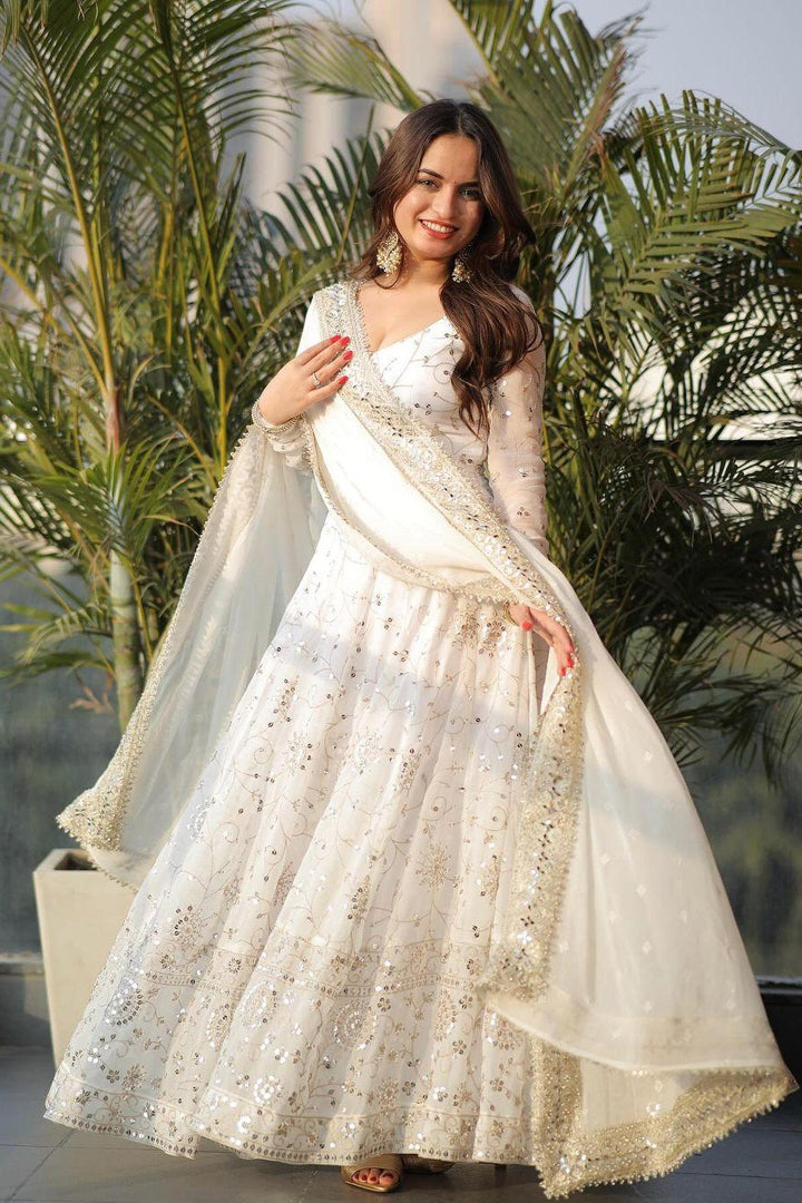Woman in a white and gold traditional outfit standing in front of green plants.