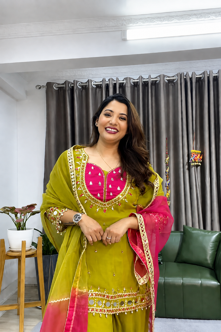 A woman modeling a traditional Indian salwar kameez suit in olive green and pink with gold embroidery, standing in a living room setting with curtains and a green sofa in the background.