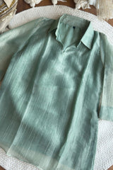 A light sage green long-sleeve collared shirt with a subtle vertical textured stripe pattern, laid flat on a white woven rug with bohemian decor in the background.
