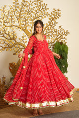 A woman modeling a red long-sleeve Anarkali gown with white floral prints and a gold border, standing in front of a decorative gold tree wall piece.