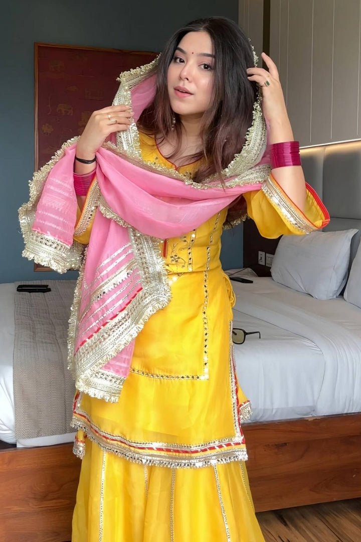 Woman in a yellow traditional outfit with a pink dupatta in a bedroom setting