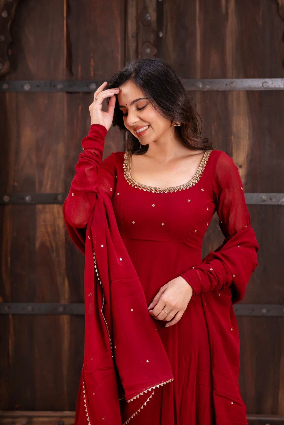 A woman wearing a deep red traditional outfit with gold embroidery and a matching dupatta, standing in front of a rustic dark wooden door.