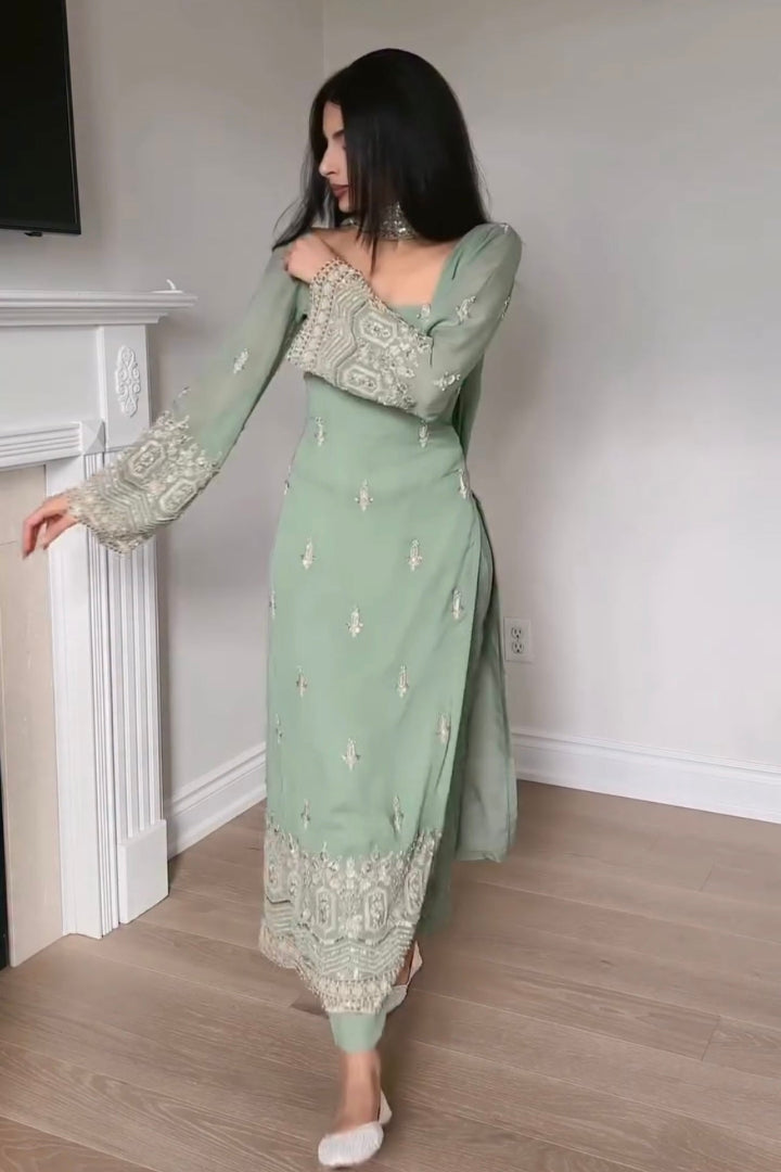 Woman wearing a green traditional outfit with intricate patterns in a room with a white fireplace.