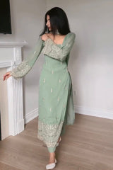 Woman wearing a green traditional outfit with intricate patterns in a room with a white fireplace.