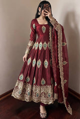 A woman models a deep maroon red anarkali gown with intricate gold, green, and blue embroidery, paired with a matching dupatta, in an indoor setting.