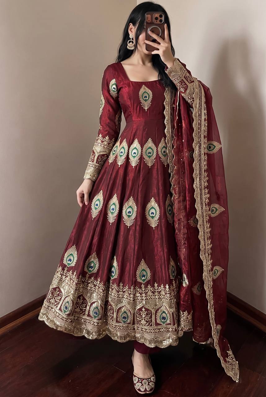 A woman models a deep maroon red anarkali gown with intricate gold, green, and blue embroidery, paired with a matching dupatta, in an indoor setting.