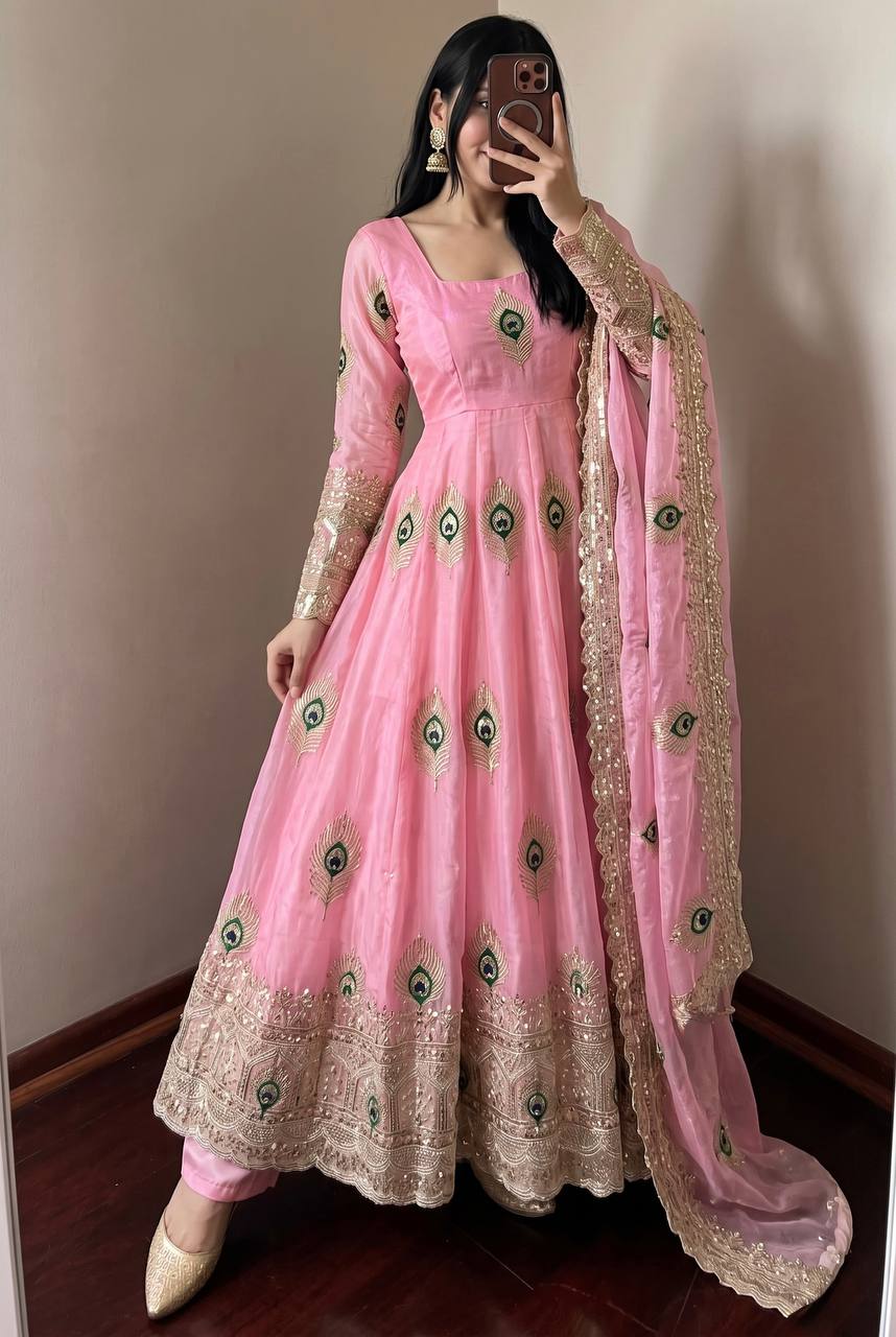 A woman wearing a pink traditional Anarkali gown with gold embroidery and green peacock motif details, paired with a matching sheer dupatta, taking a mirror selfie in an indoor setting.