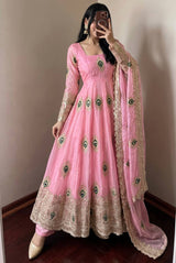 A woman wearing a pink traditional Anarkali gown with gold embroidery and green peacock motif details, paired with a matching sheer dupatta, taking a mirror selfie in an indoor setting.