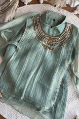 A sage green long-sleeve kurta with vertical stripes and gold metallic embroidery around the high neckline, laid flat on a textured white woven rug.