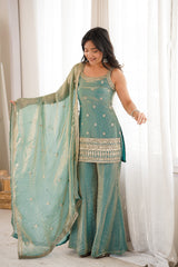 Woman in a teal traditional outfit standing in a bright room with white curtains.