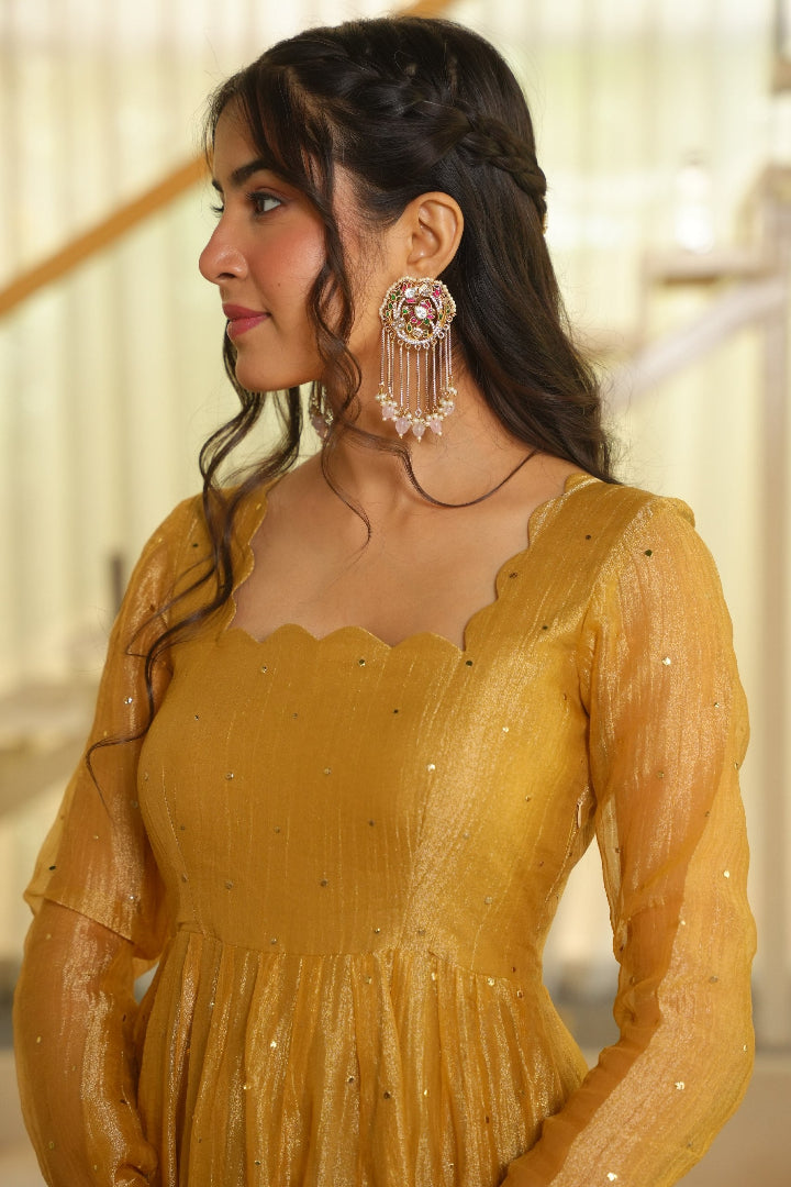 A woman wearing a mustard yellow embroidered dress and matching long tassel earrings, shown in profile against a softly blurred indoor background.