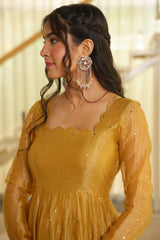 A woman wearing a mustard yellow embroidered dress and matching long tassel earrings, shown in profile against a softly blurred indoor background.
