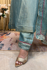 A close-up of a person wearing a teal-green traditional outfit with matching pants and a sheer embroidered dupatta, paired with metallic gold strappy sandals. The setting is indoors with a patterned rug and curtains in the background.