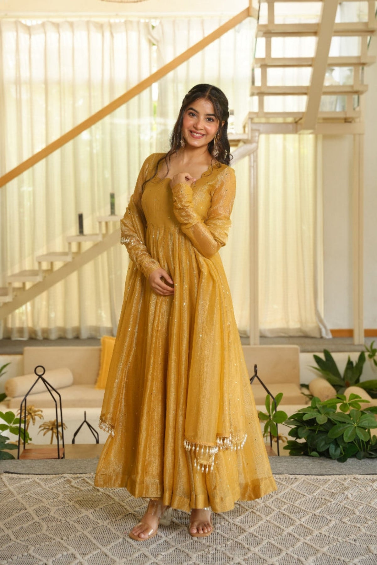 A woman stands barefoot in a bright, modern indoor space wearing a long mustard yellow embroidered dress with sheer sleeves and decorative beadwork near the hem.