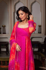 Woman in a vibrant pink and orange traditional outfit with intricate patterns, standing in an elegant indoor setting.