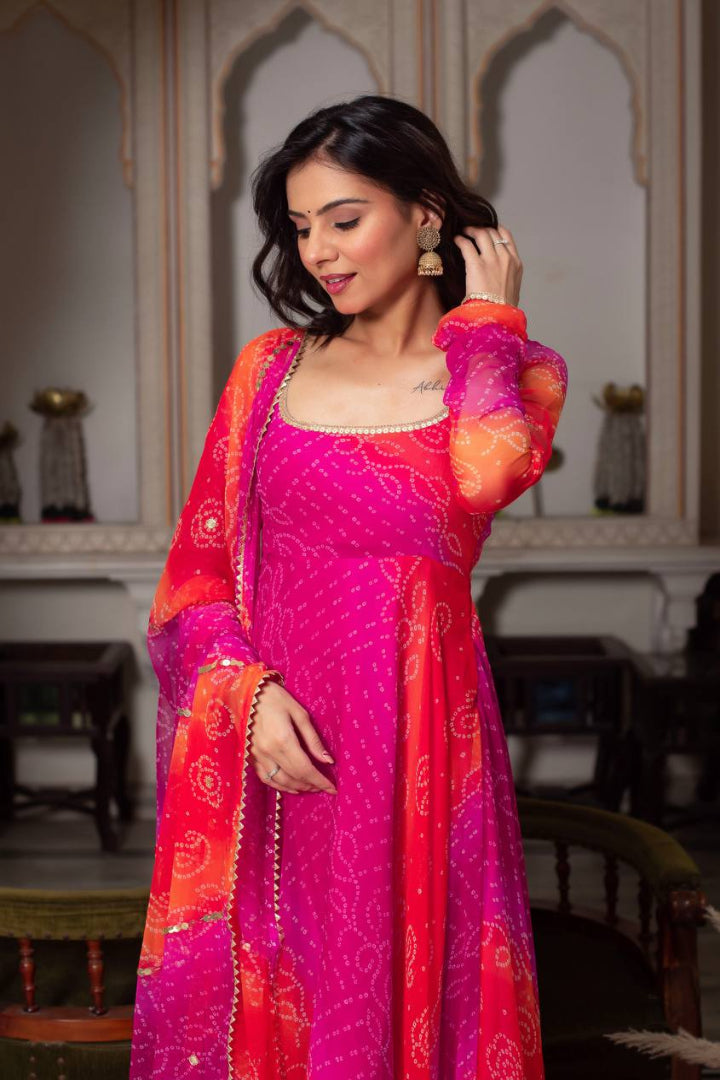 Woman in a vibrant pink and orange traditional outfit with intricate patterns, standing in an elegant indoor setting.