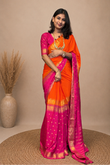 A woman modeling a bright orange and magenta pink saree with gold borders and polka dots, standing on a round woven mat against a beige studio backdrop.