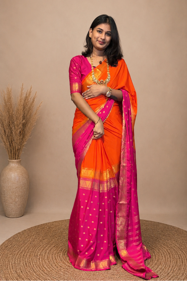 A woman modeling a bright orange and magenta pink saree with gold borders and polka dots, standing on a round woven mat against a beige studio backdrop.