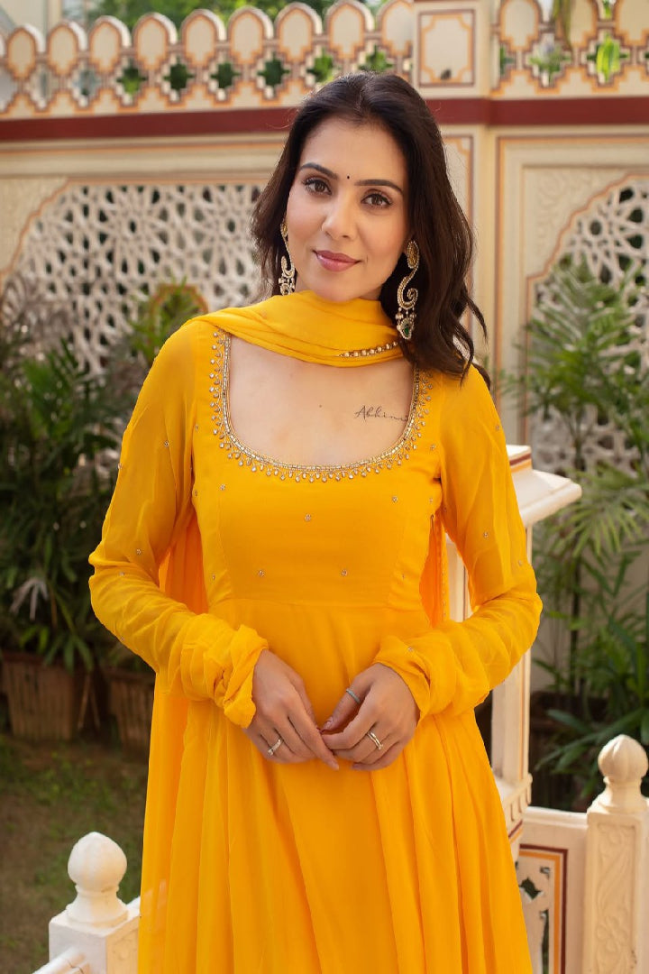 Woman in a yellow dress with intricate detailing standing in front of decorative wall and plants.