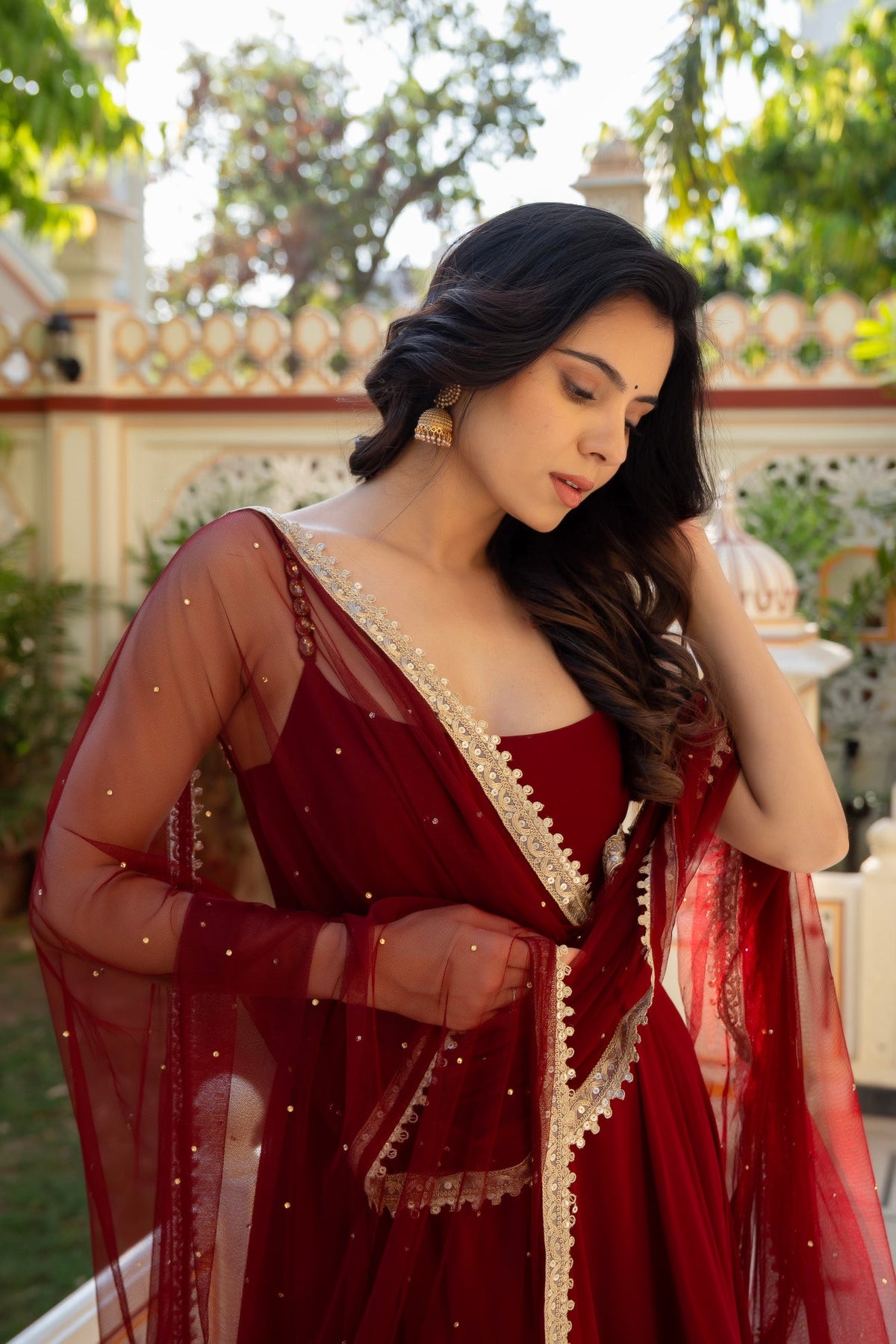 A woman wearing a deep red traditional dress with a sheer embroidered drape, standing outdoors in front of a decorative white wall and greenery.