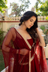 A woman wearing a deep red traditional dress with a sheer embroidered drape, standing outdoors in front of a decorative white wall and greenery.