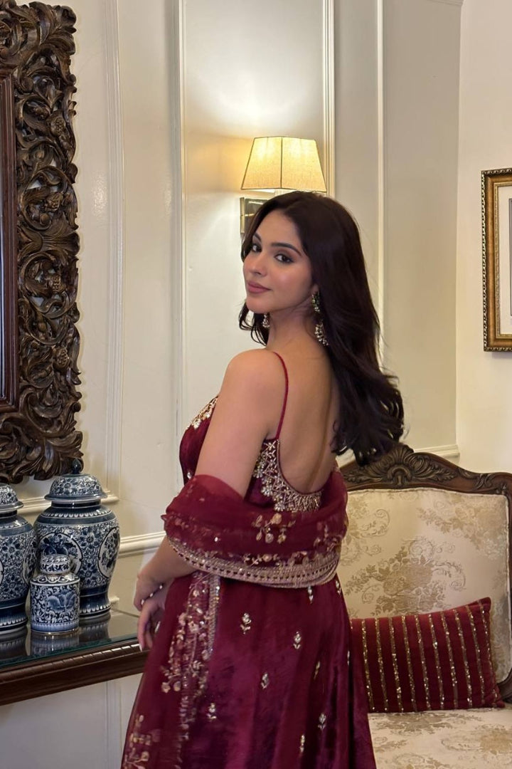 Woman in a maroon traditional outfit standing in a decorated room with a mirror and lamp.