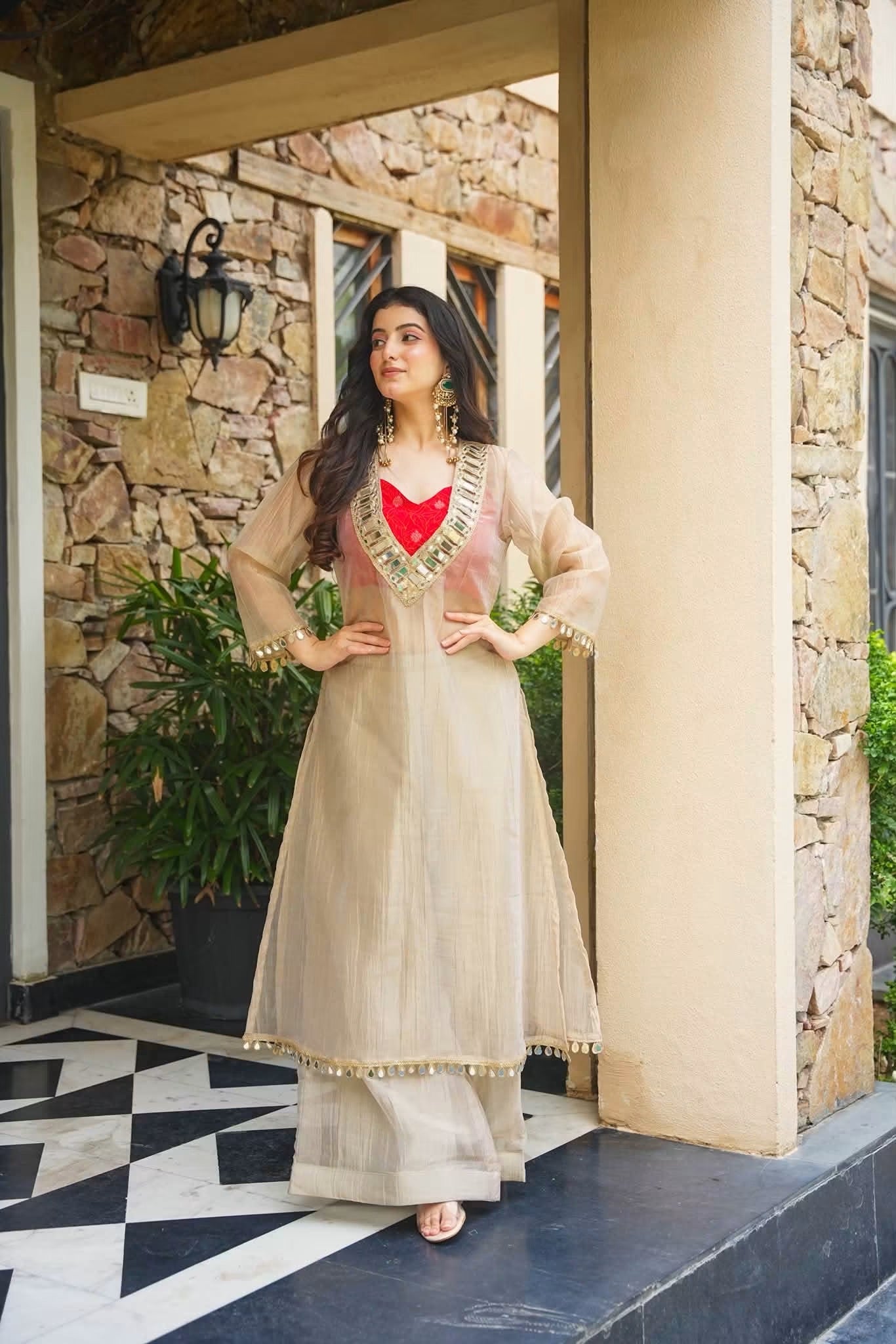 A woman models a beige traditional Indian outfit with a red heart-shaped neckline accent and gold embroidery, standing on a stone porch with a black and white tiled floor.