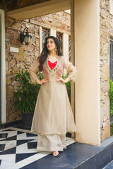 A woman models a beige traditional Indian outfit with a red heart-shaped neckline accent and gold embroidery, standing on a stone porch with a black and white tiled floor.