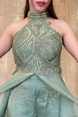 A close-up of a model wearing a metallic sage green halter jumpsuit with intricate silver beadwork on the bodice and matching sheer draped fabric at the waist, set against a beige patterned wall.