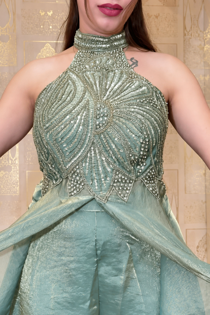 A close-up of a model wearing a metallic sage green halter jumpsuit with intricate silver beadwork on the bodice and matching sheer draped fabric at the waist, set against a beige patterned wall.