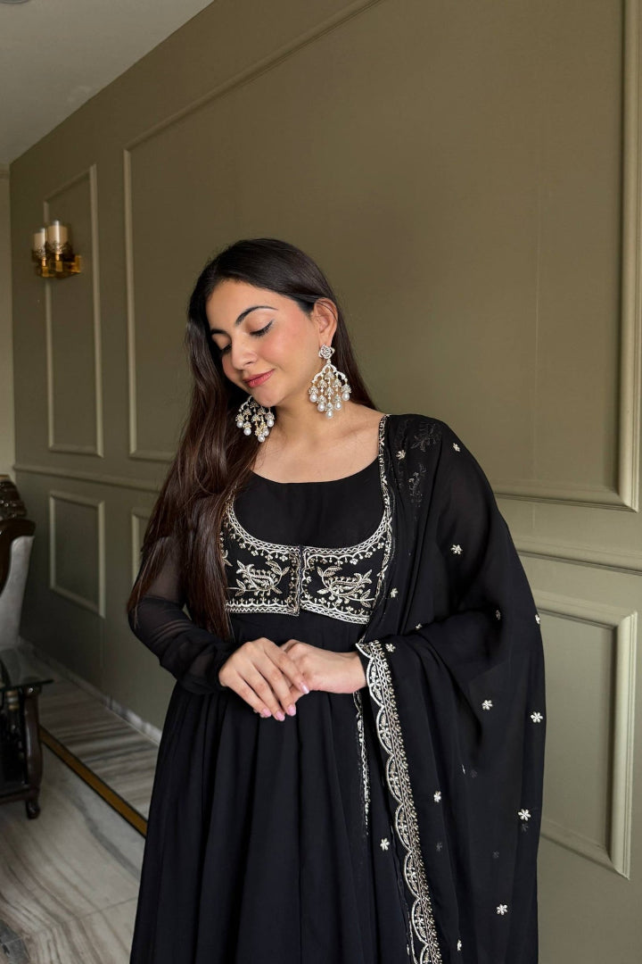 Woman in a black dress with silver embroidery standing in a room with a neutral wall.