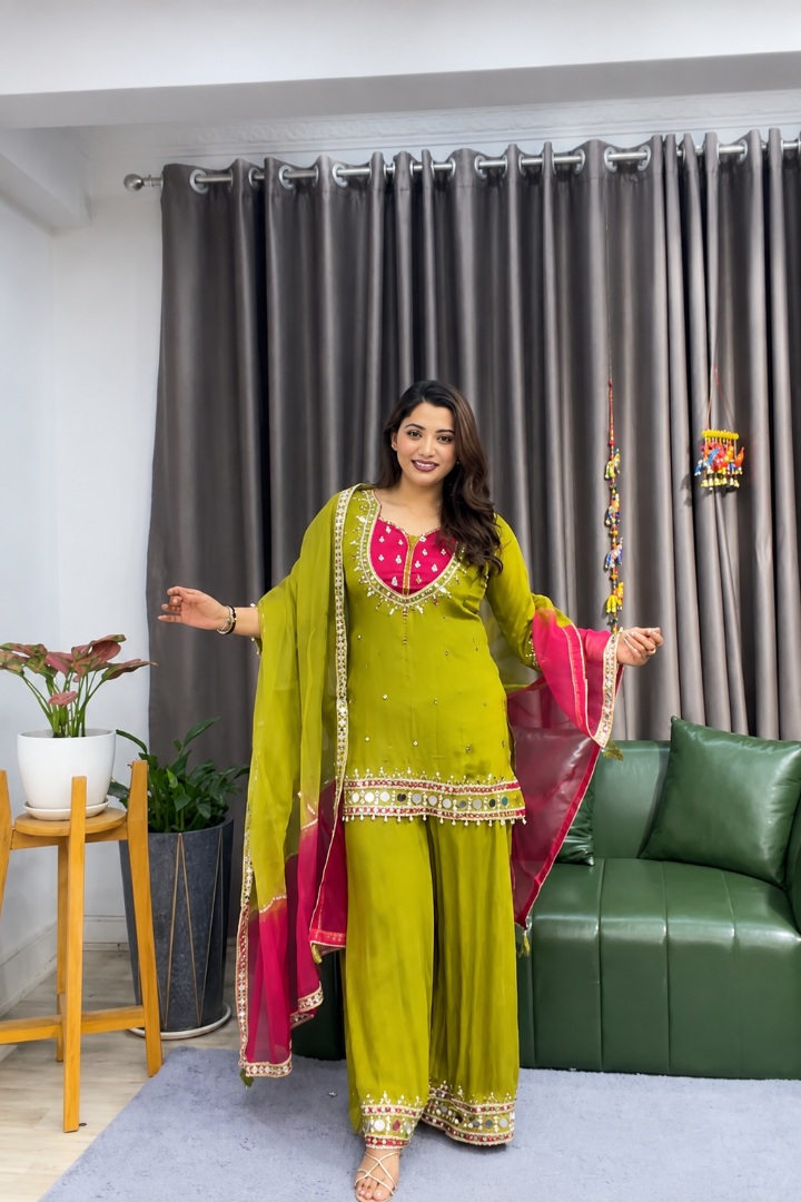 A woman modeling a traditional Indian outfit consisting of an olive green kurta and wide-leg pants with a bright pink dupatta, standing in a living room setting with a green sofa and gray curtains.
