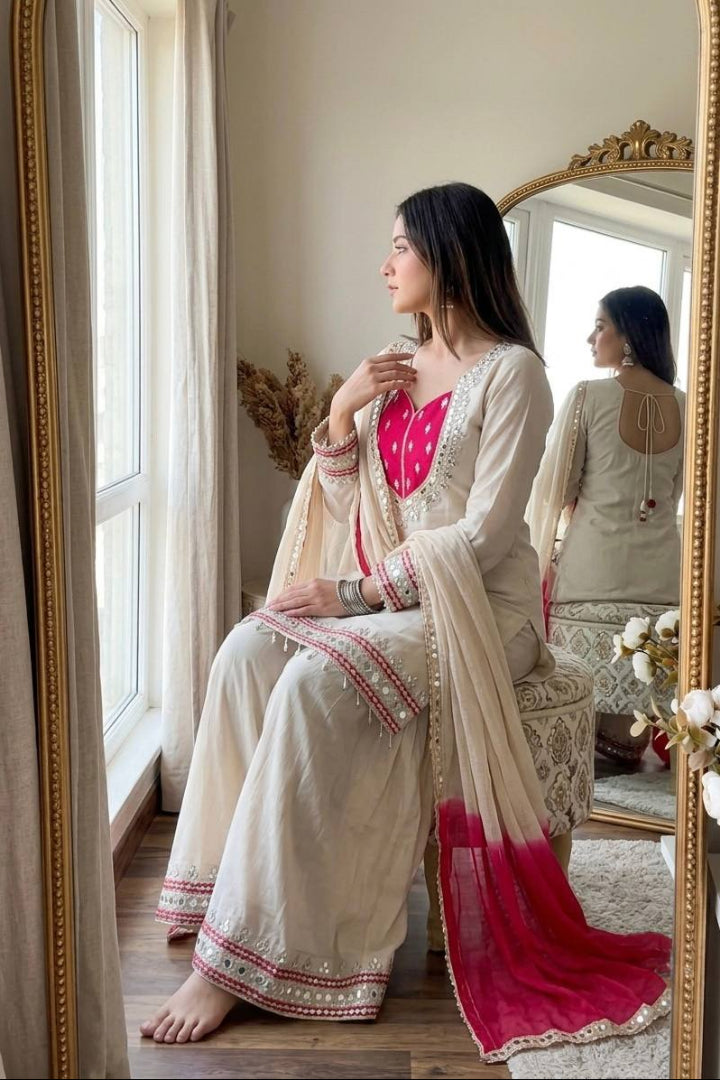 A woman sits barefoot in front of a window, wearing a traditional cream-colored outfit with bright pink embroidery and a matching dupatta with a red-to-pink gradient. She is seated on a patterned stool, with a large gold-framed mirror behind her reflecting her back.
