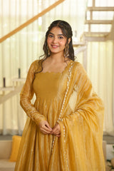 A woman wearing a mustard yellow traditional dress with a matching sheer dupatta draped over her shoulders, standing in a brightly lit indoor setting with a staircase in the background.