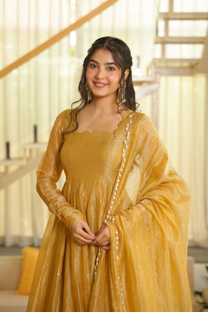 A woman wearing a mustard yellow traditional dress with a matching sheer dupatta draped over her shoulders, standing in a brightly lit indoor setting with a staircase in the background.
