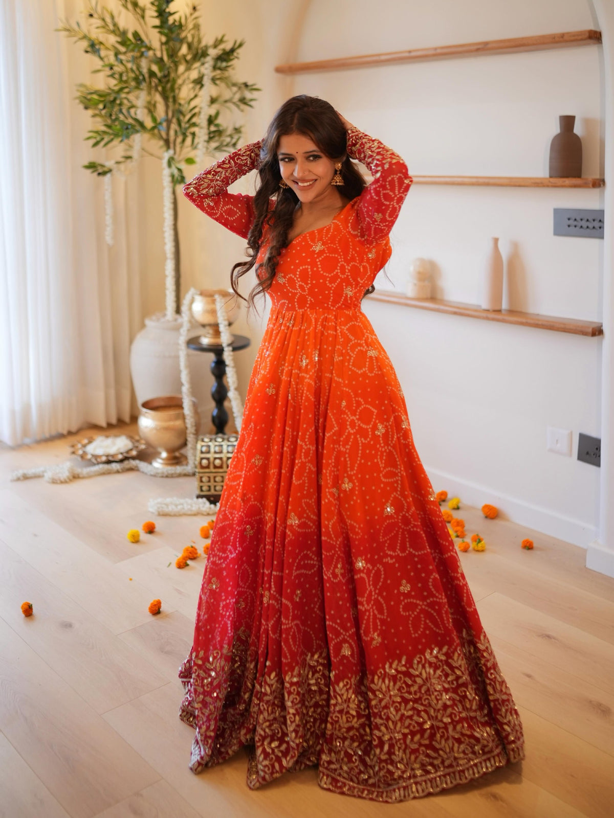 A woman wearing a vibrant orange and red embroidered Anarkali gown with gold detailing, posing in a bright, modern room with wooden floors and shelves.