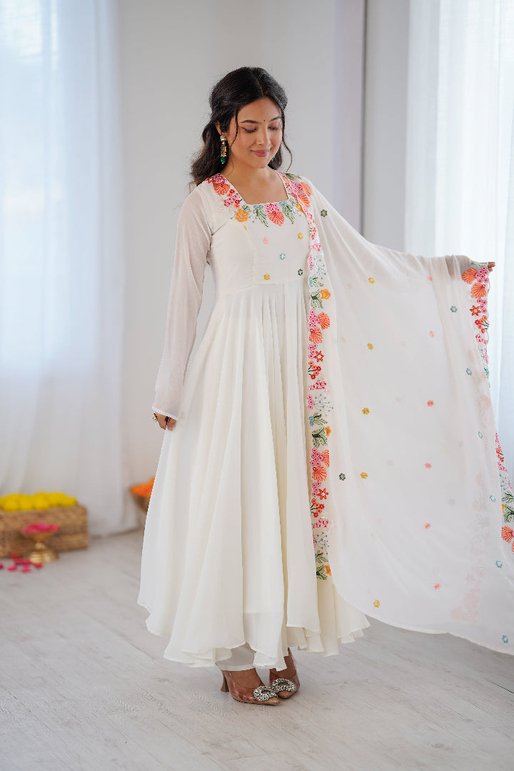 A woman modeling a white long-sleeve Anarkali gown with colorful floral embroidery around the neckline and a matching sheer dupatta draped over her arm, set in a bright, minimalist room with white curtains.