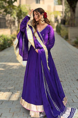 Woman in a traditional purple and gold outfit standing on a paved path with greenery in the background.